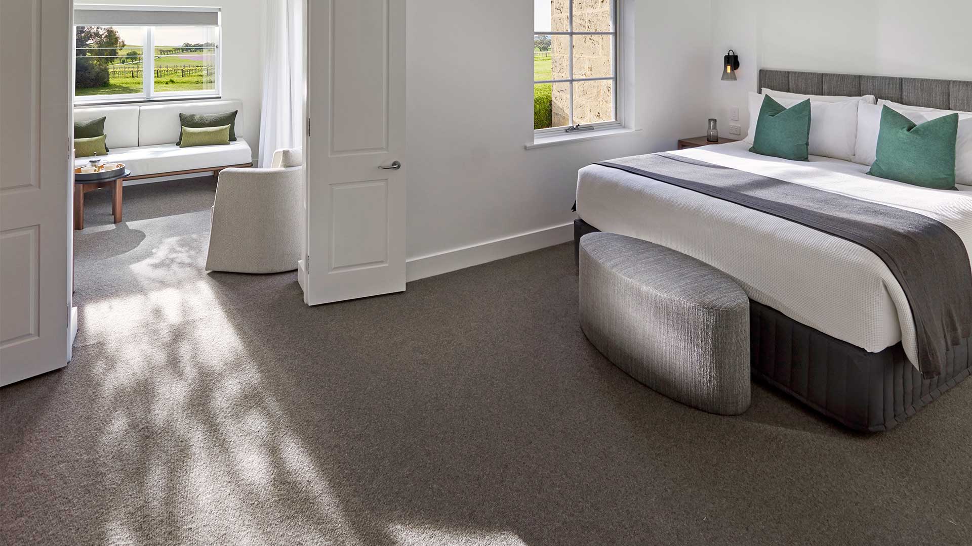 A guest bedroom with a white linen bed, deep green cushions and a curved grey ottoman, standing on wall-to-wall taupe wool carpet with dappled tree shadow falling across it, a window seat and open paddocks beyond