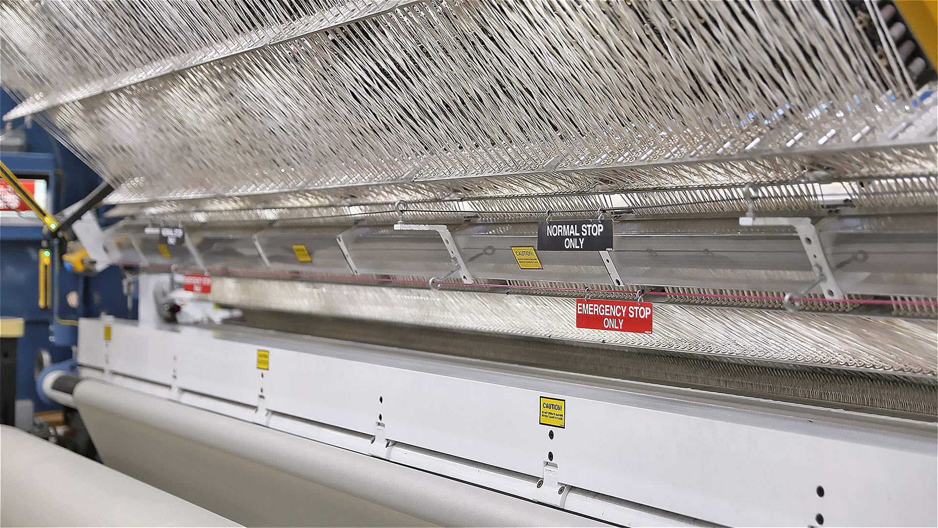 A wide tufting machine on the factory floor, a long row of needles threaded with pale yarn running the length of the frame above the backing cloth, with Normal Stop and Emergency Stop signs hanging from the guard rail