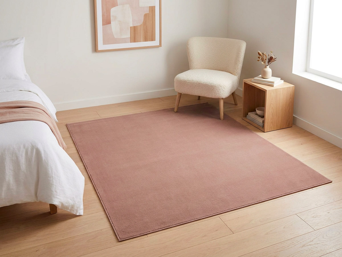 A square dusty pink rug on a timber floor in a bedroom, beside a white bed, a cream boucle chair and an oak side table