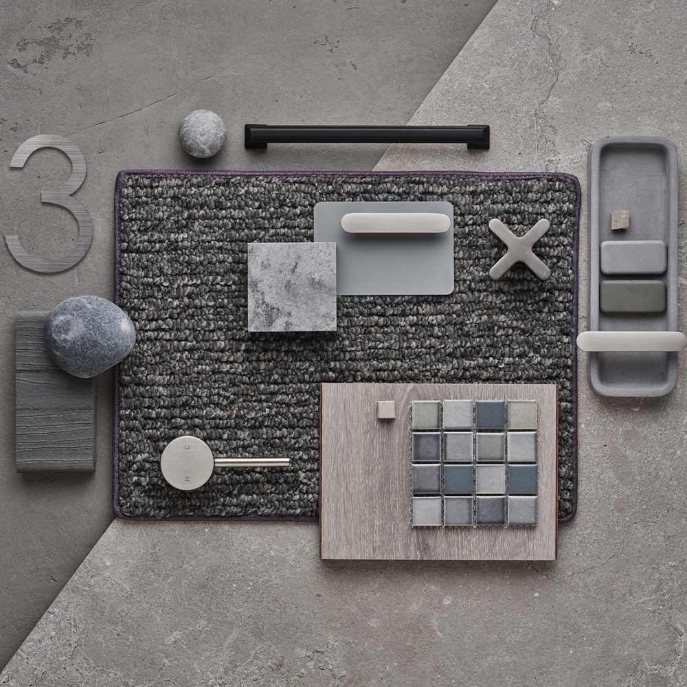 An overhead flat-lay of a charcoal loop-pile rug on a polished concrete floor, laid out with grey stone and marble offcuts, a limed timber board, a square of blue-grey mosaic tile, a felted wool pebble and a brushed metal tap